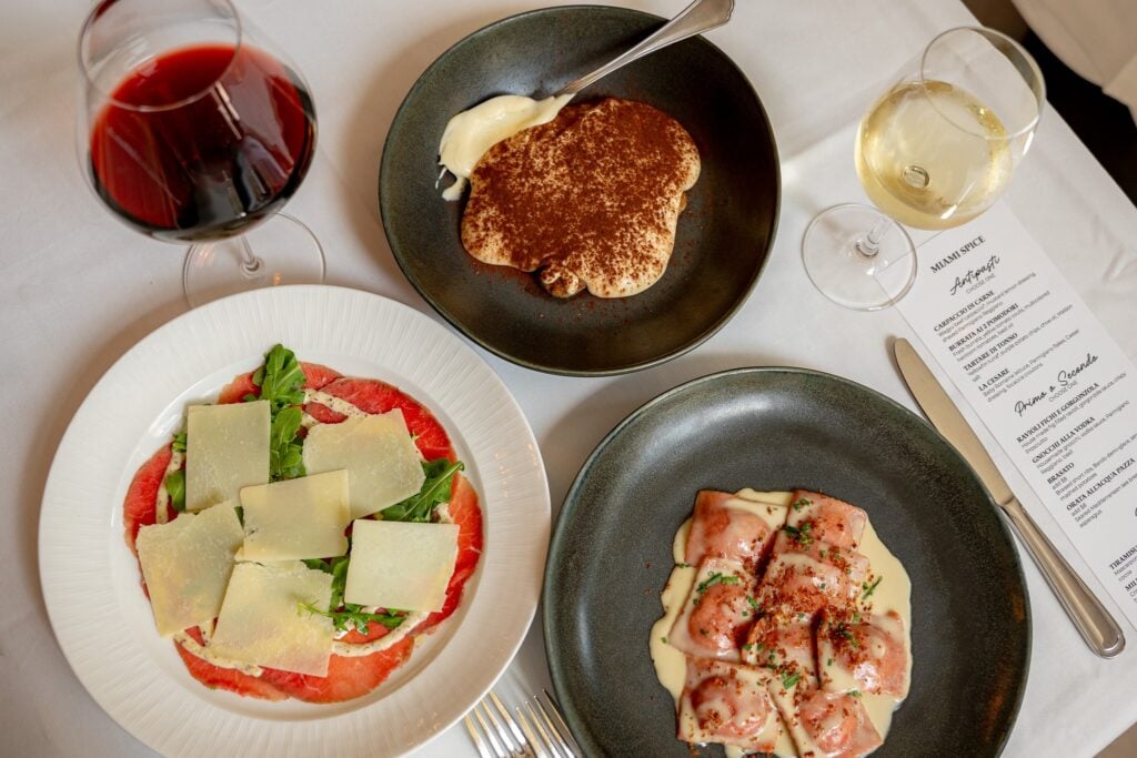 A Miami Spice spread at Da Angelino Cucina Italiana with beef carpaccio, ravioli fichi e gorgonzola, tiramisu, and glasses of red and white wine beside the printed menu.