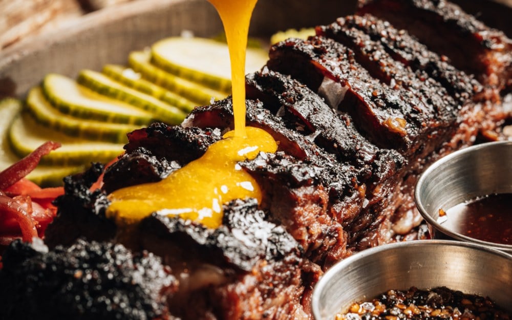 Wood-fired short rib at KYU being finished with a pour of golden sauce, served with house pickles and dipping sauces.