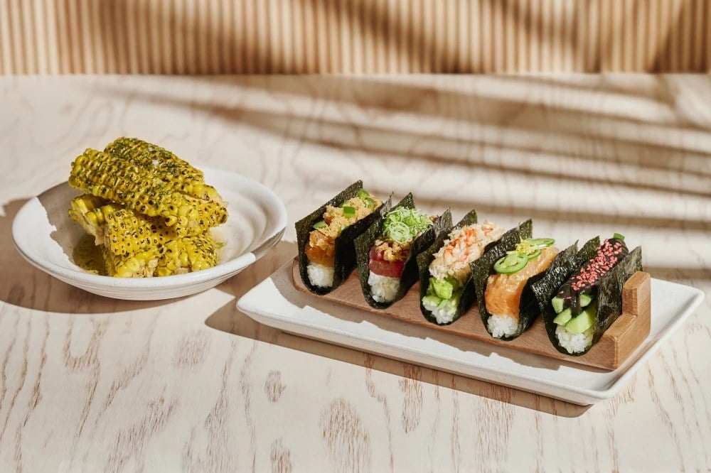 A row of five open-style hand rolls on a wooden stand at Nami Nori, served alongside a bowl of charred corn.