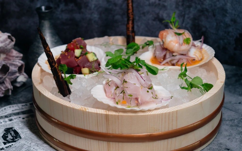 A ceviche sampler at Paperfish Sushi served on scallop shells over crushed ice in a wooden hangiri, with tuna, shrimp, and white fish.
