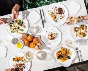 An overhead view of a sunlit table at Le Jardinier Miami covered with plated dishes, including branzino, steak, beef tartare, shrimp, seafood stew, a basket of bread, and glasses of white wine.
