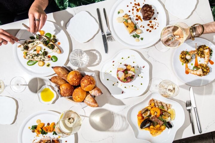 An overhead view of a sunlit table at Le Jardinier Miami covered with plated dishes, including branzino, steak, beef tartare, shrimp, seafood stew, a basket of bread, and glasses of white wine.