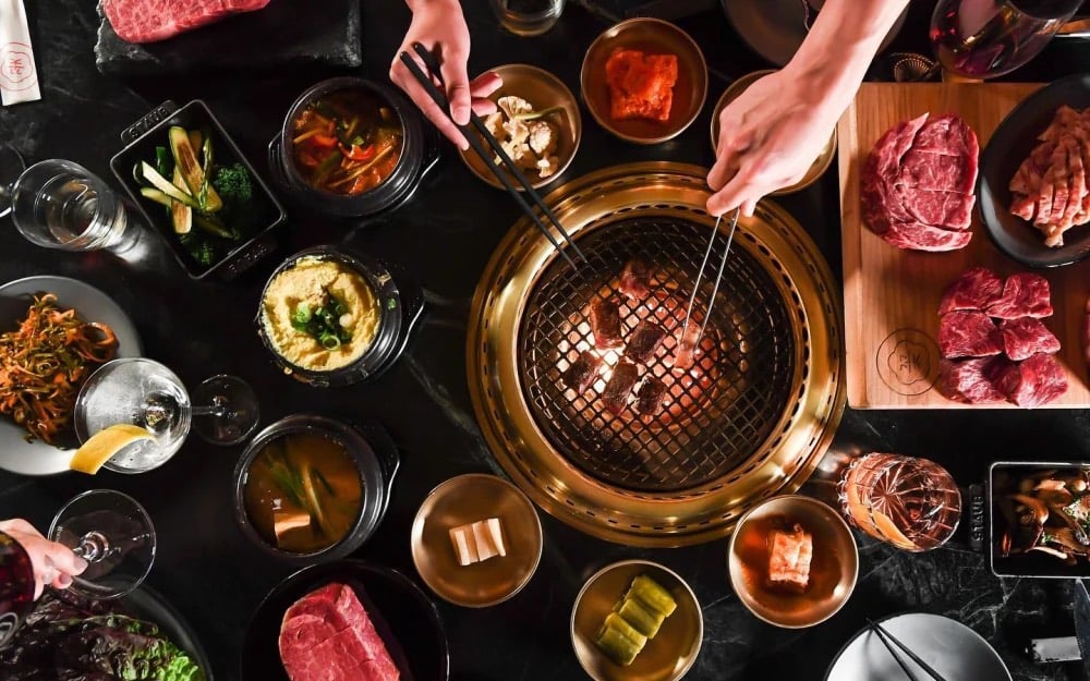 An overhead view of a table at COTE Miami with a built-in grill searing beef in the center, surrounded by raw cuts of dry-aged steak on a wooden board, banchan, stews, and cocktails.