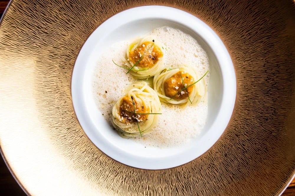 Three nests of thin pasta topped with a golden sauce and chives, served in a foamed broth in a white bowl set on a textured gold charger at L'Atelier de Joël Robuchon.