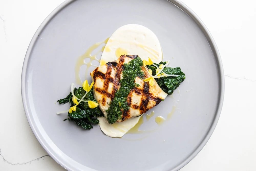 A grill-marked fillet of fish topped with green herb sauce, served over sautéed kale with a swipe of pale purée on a gray plate at Le Jardinier Miami.
