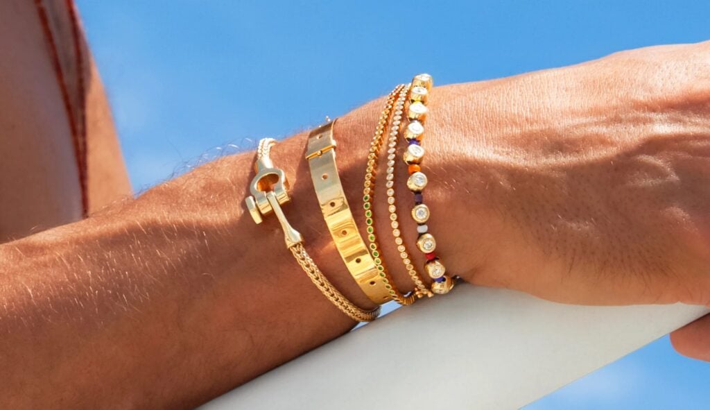 Stacked gold Luis Morais bracelets and bangles worn on a wrist against a blue sky