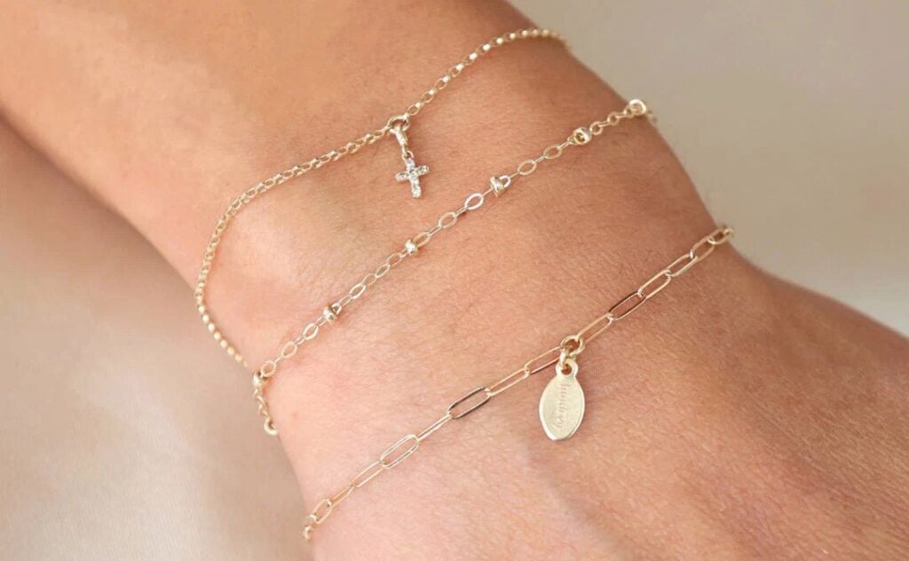 Delicate gold Taudrey chain bracelets layered on a wrist with a small charm and cross pendant