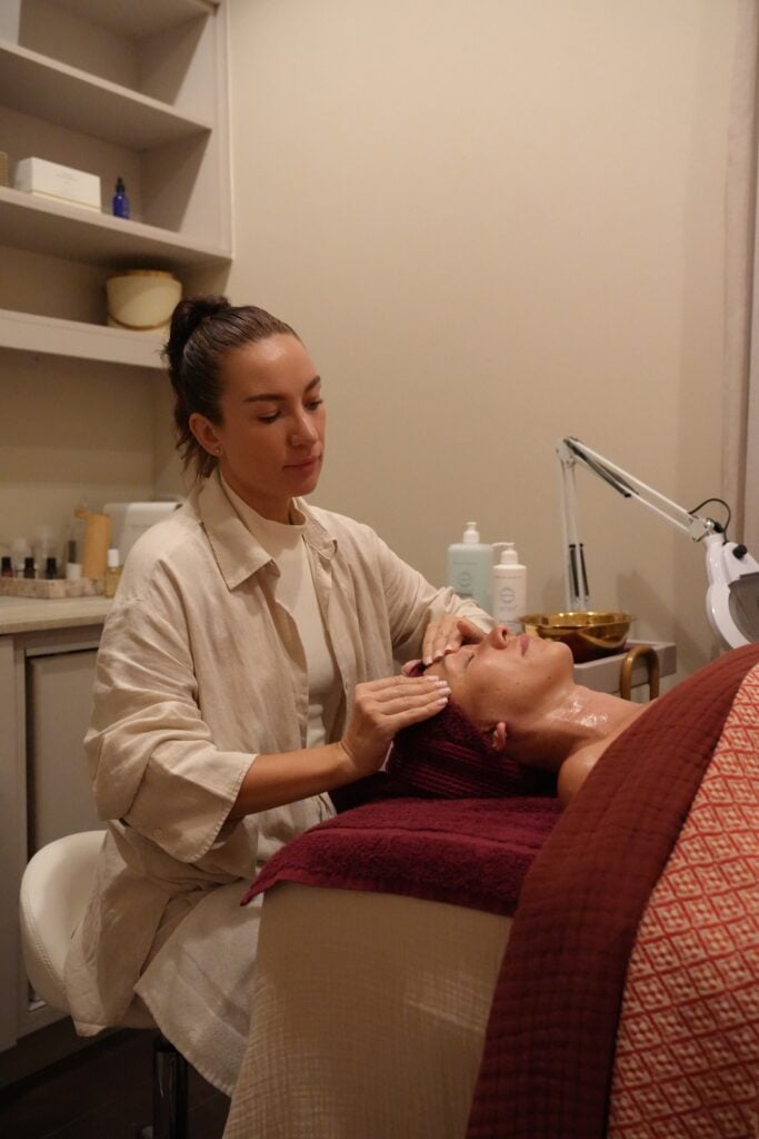 An esthetician works on a client's face in a treatment room at Da Beauty Bar, with burgundy towels and a magnifying lamp