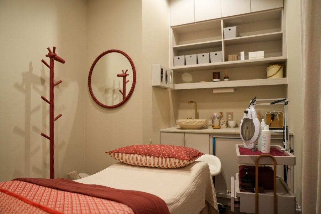 A treatment room at Da Beauty Bar with a draped bed, terracotta throw, round red mirror, and open shelving